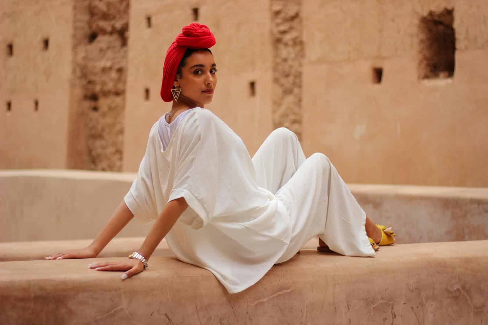 Stylish woman in white outfit with red headscarf poses outdoors in Marrakesh courtyard.