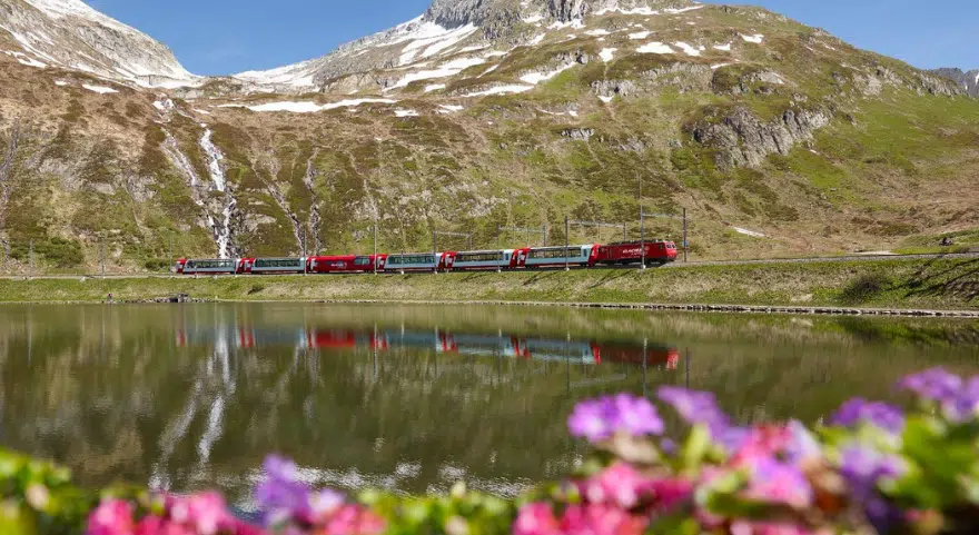 The Glacier Express travels along a lake near the Oberalp Pass, with snow-streaked mountains reflected in the still water.
