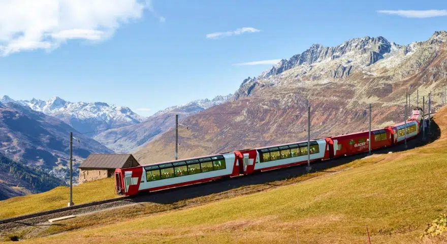 The Glacier Express curves along a hillside above a Swiss alpine valley, with snow-capped peaks in the distance.