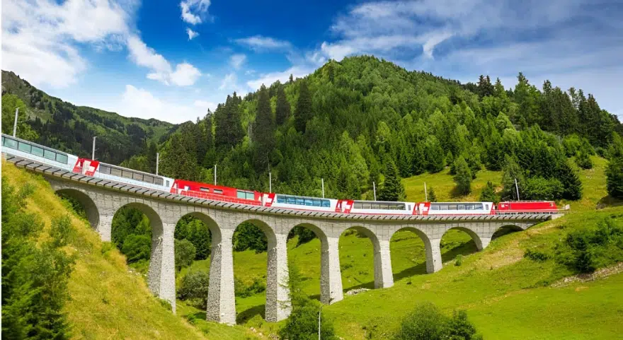 The Glacier Express crosses the curved stone arches of the Landwasser Viaduct surrounded by forested Swiss hillsides.