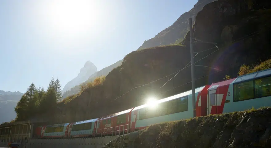 The Glacier Express Switzerland route passes below a rocky mountainside near Zermatt with the Matterhorn visible in the distance.