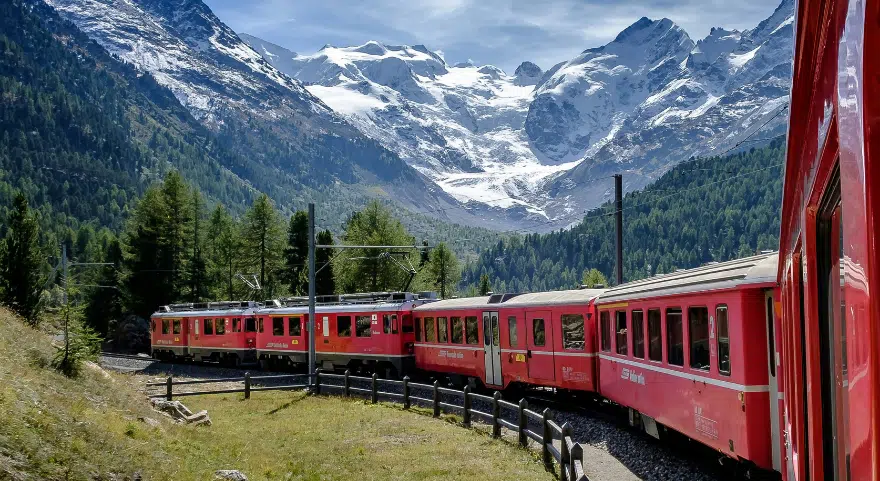 The Bernina Express curves through a valley with snow-covered peaks and a glacier in the background.