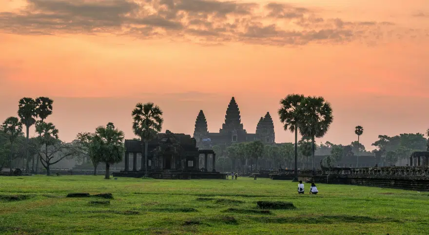 Angkor Wat sunrise silhouettes the temple's spires against an orange sky, reflected in the still water of the ponds.