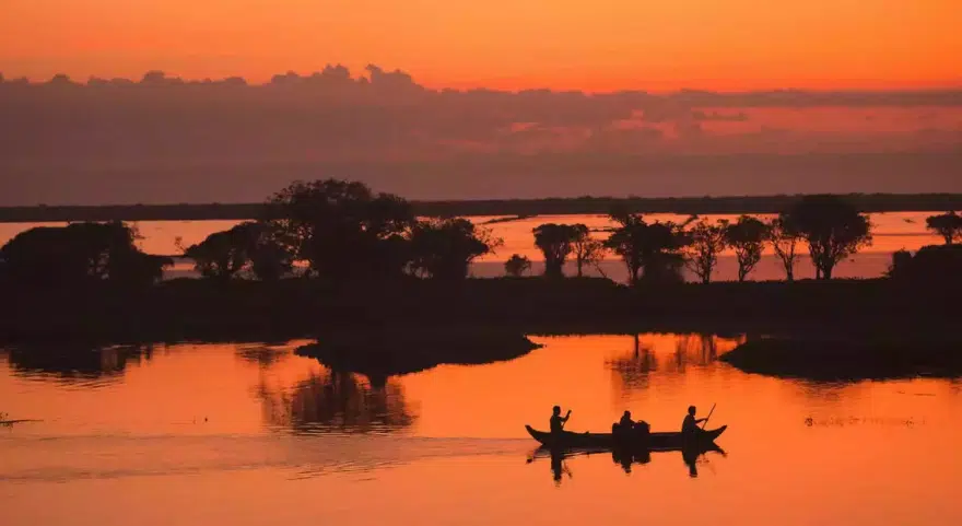 A small boat crosses Tonlé Sap at sunset, silhouetted against an orange sky reflected in the still water.