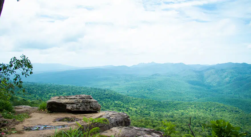 A forested valley in Cambodia seen from a rocky hilltop viewpoint.