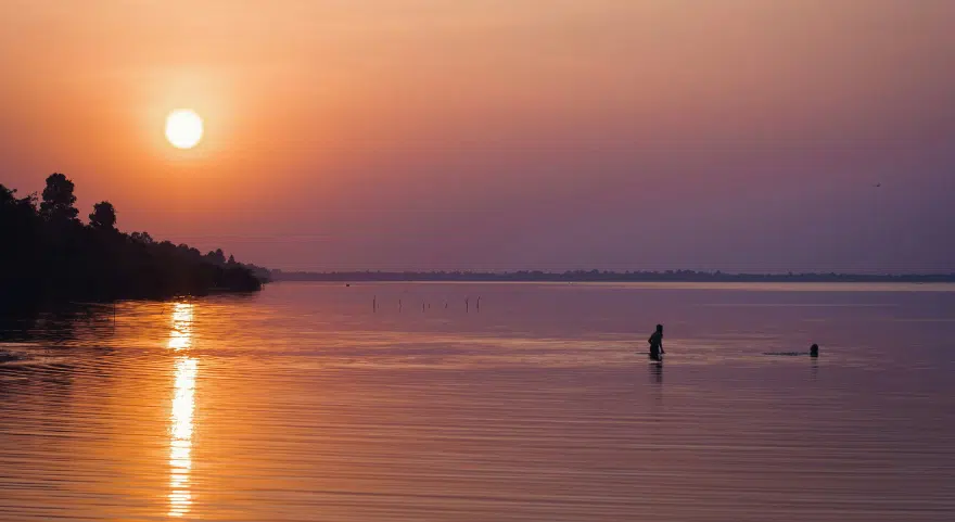 The sun sets over Tonlé Sap Lake, casting orange light across the still water near the shore.
