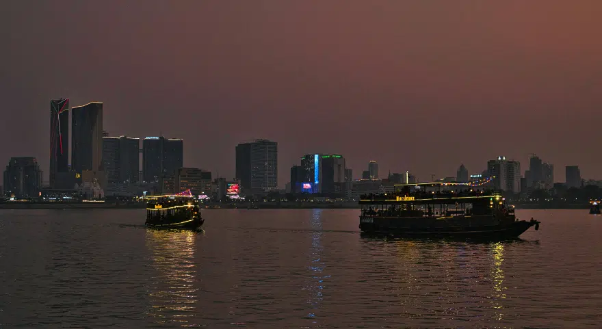 Boats cross the Mekong River at dusk with the Phnom Penh skyline lit up in the background.